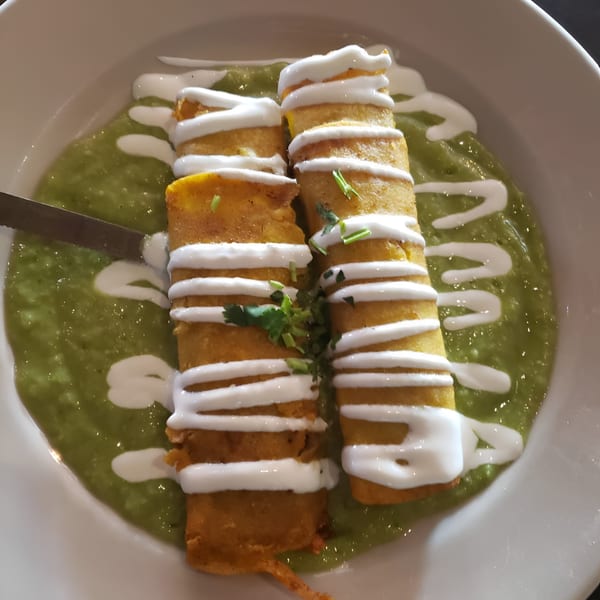 Two golden taquitos drizzled with white cream, resting in a vibrant green sauce, garnished with cilantro.