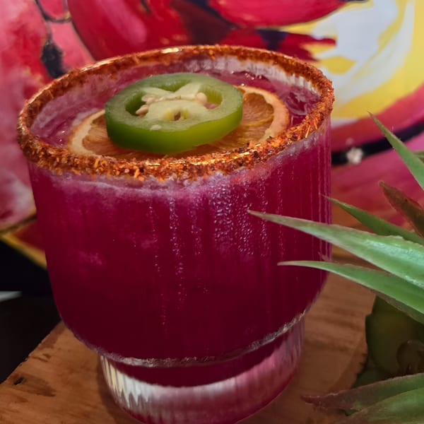 Vibrant red margarita with a chili-lime rim, orange slice, and jalapeño garnish, on a wooden surface.